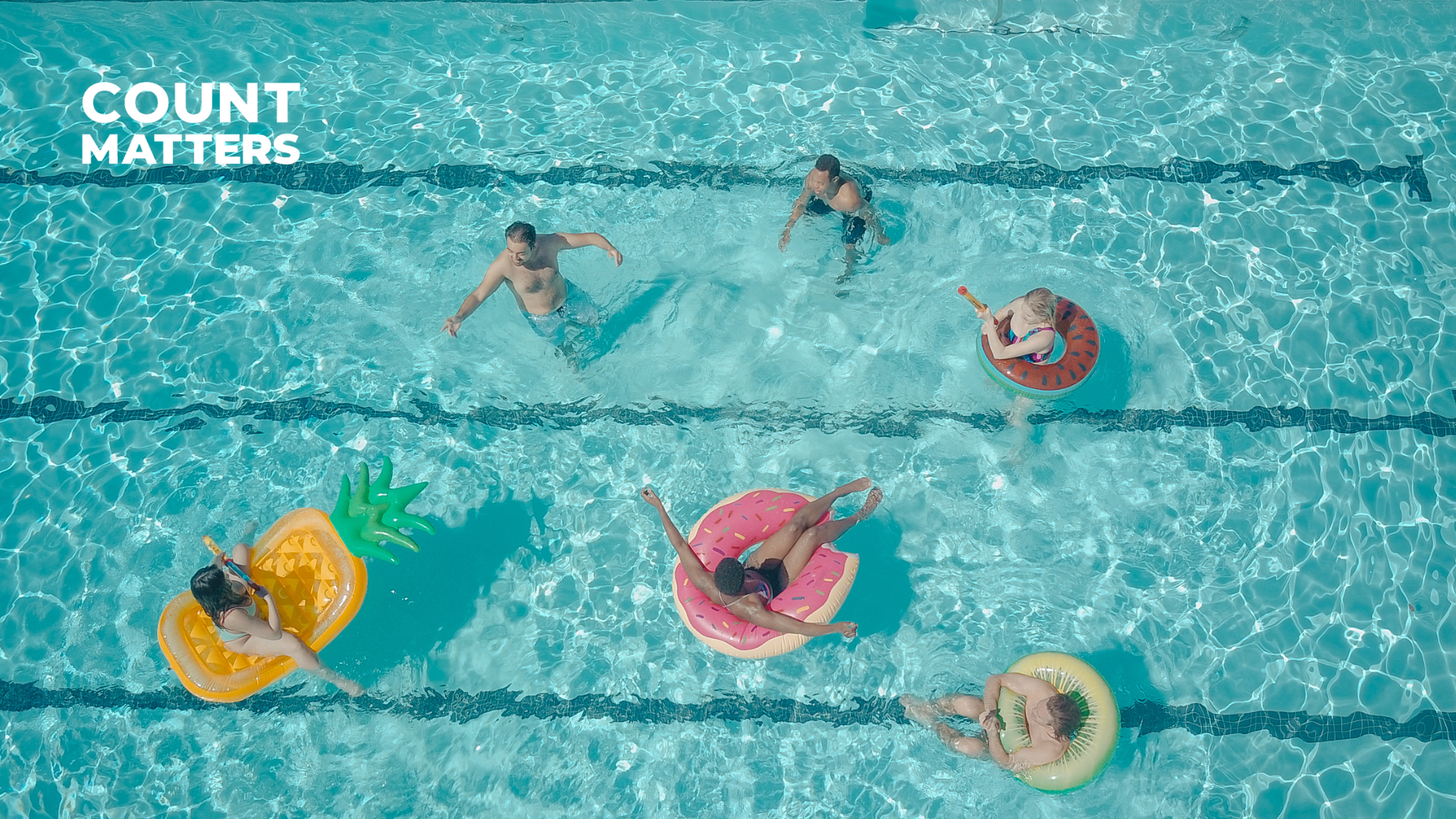 Luftfoto af en swimmingpool med personer, der nyder det på farverige oppustelige baderinge, herunder ananas-, donut- og vandmelon-design, med CountMatters-logoet i øverste venstre hjørne