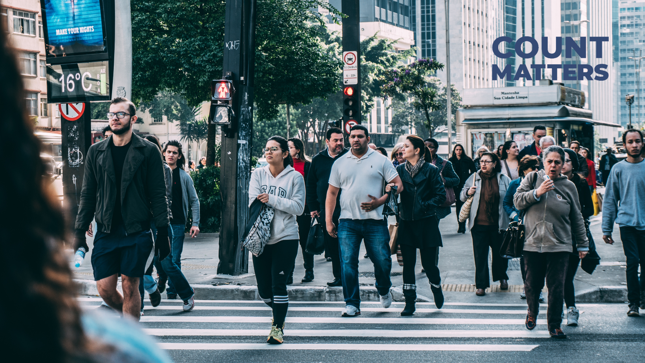 A busy pedestrian crosswalk in an urban setting with diverse individuals walking in different directions. The background features modern high-rise buildings, trees, and city signage, including a digital temperature display. The 'CountMatters' logo is visible in the top right corner, emphasizing visitor analytics and foot traffic monitoring in smart cities.