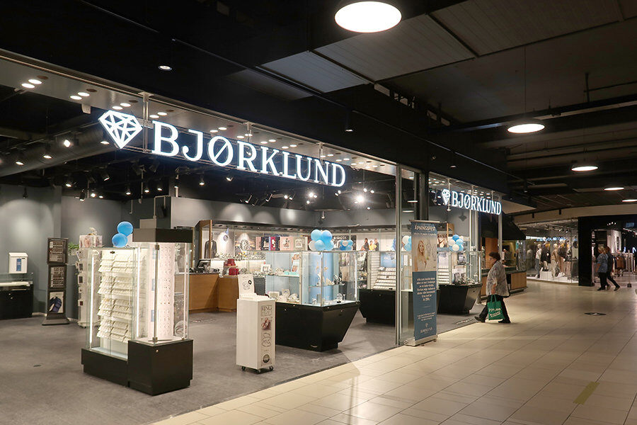 Bjørklund-butik med upplyst skylt i en galleria, visar utställda smycken och klockor i glasmontrar. Besökare promenerar förbi.